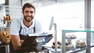 Bakery owner leaning against the till system with bread and cakes displayed in the background