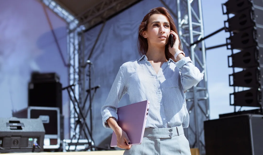 Woman holding a laptop and speaking on the phone at an outdoor event stage setup, with rigging and speakers visible behind her