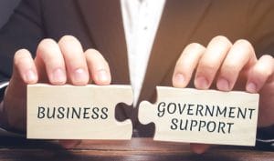 Hands holding wooden jigsaw puzzle pieces with the words ‘Business’ and ‘Government Support’ fitting together, symbolising financial support for companies.