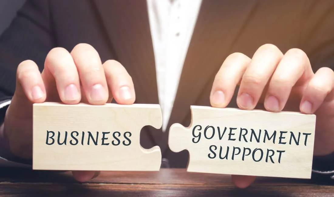 Hands holding wooden jigsaw puzzle pieces with the words ‘Business’ and ‘Government Support’ fitting together, symbolising financial support for companies.