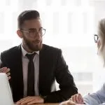 Man in a suit talking to a colleague at a desk with a laptop, discussing financial matters like commission disclosure