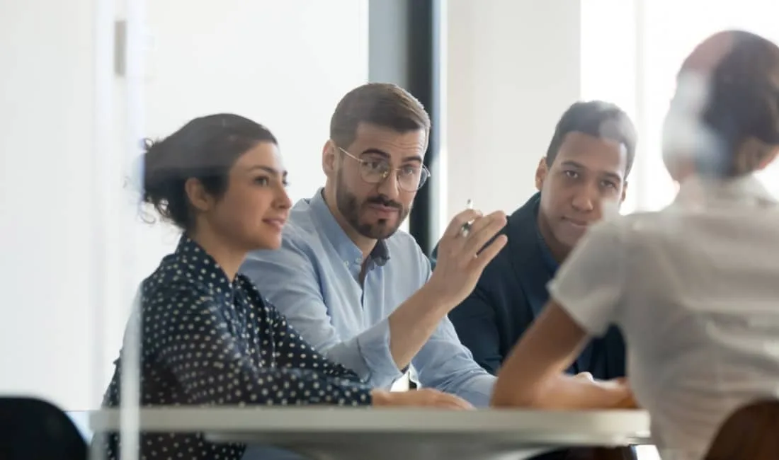 Three professionals giving commission disclosure in a meeting room listening to a colleague, with one man gesturing while speaking