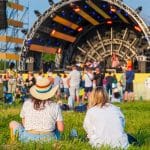 Crowd sitting and standing in a grassy field watching a live outdoor stage performance during the day