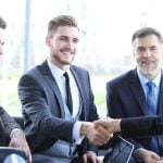 Four professionals sat down whilst two of them shake hands, smiling at one another.