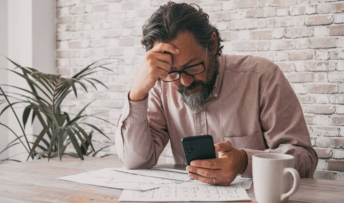 Stressed businessman looking at his phone with financial documents on the table, representing the risks of personal guarantees without protection.