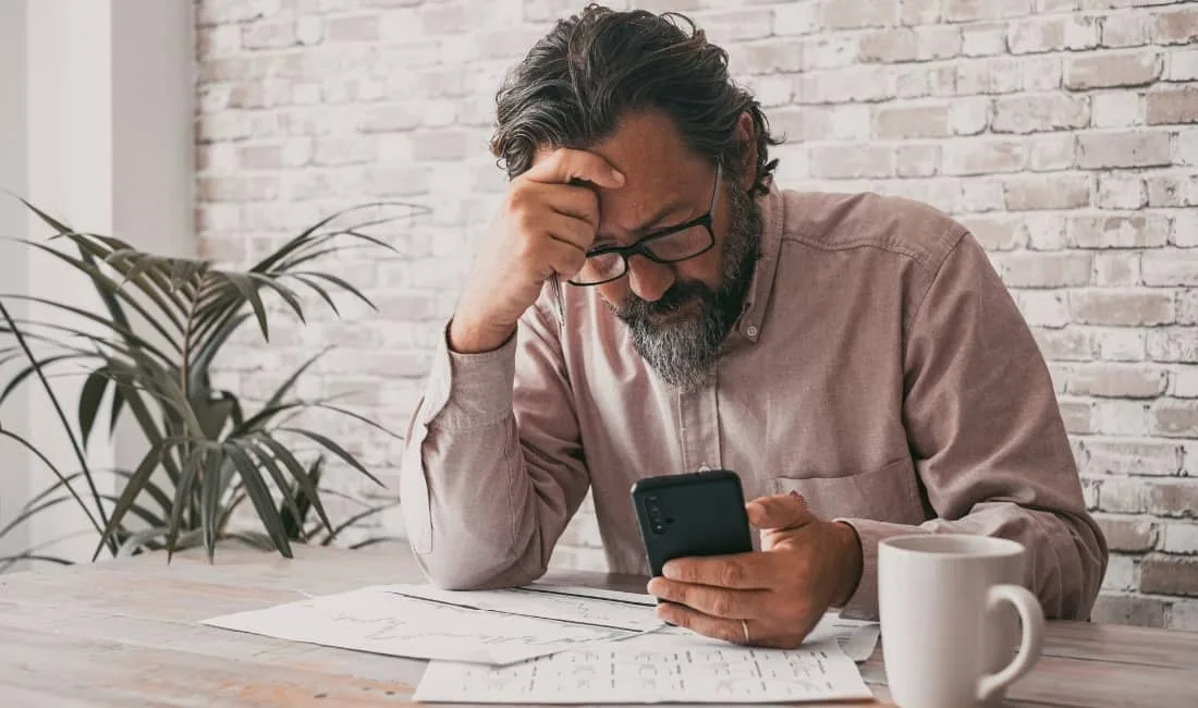 Stressed businessman looking at his phone with financial documents on the table, representing the risks of personal guarantees without protection.