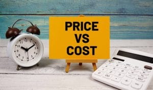 Sign reading "PRICE VS COST" sales tactic placed between a clock and a calculator on a wooden surface