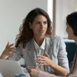 Two women in conversation at a desk with a laptop, one explaining customer motivation with hand gestures