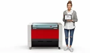 Woman standing next to a red and grey Trotec Speedy 400 laser machine, holding a tablet displaying a design interface