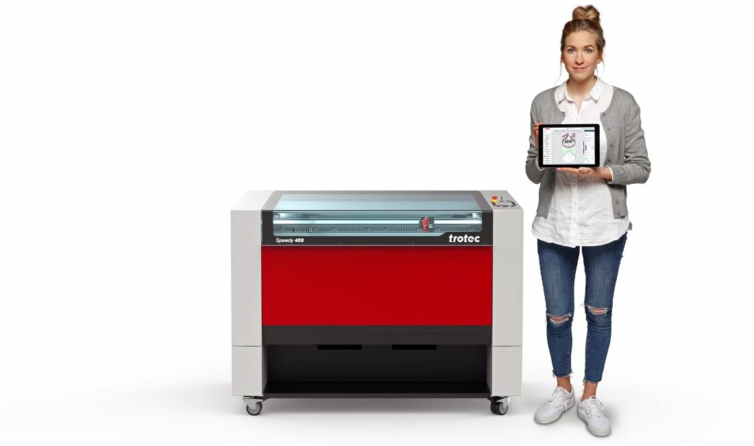 Woman standing next to a red and grey Trotec Speedy 400 laser machine, holding a tablet displaying a design interface